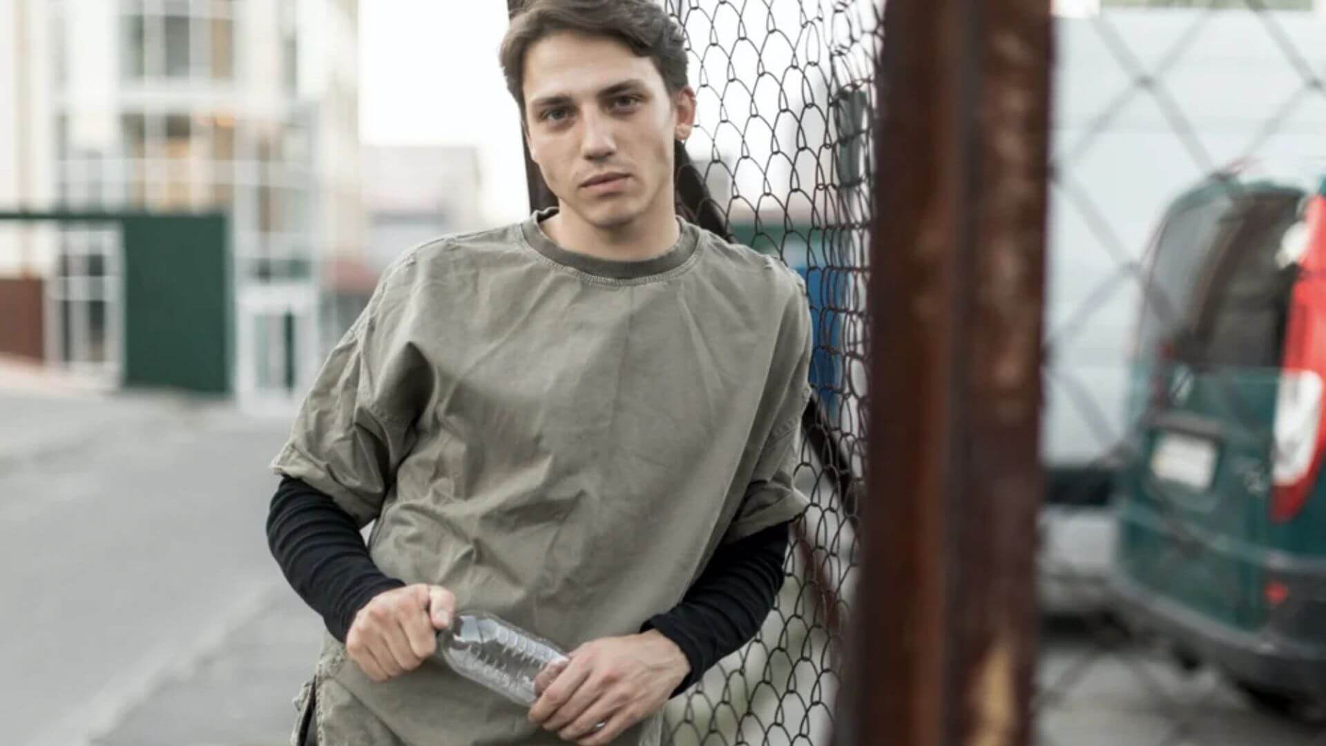 A young man leans against a wooden fence, holding a bottle in his right hand.