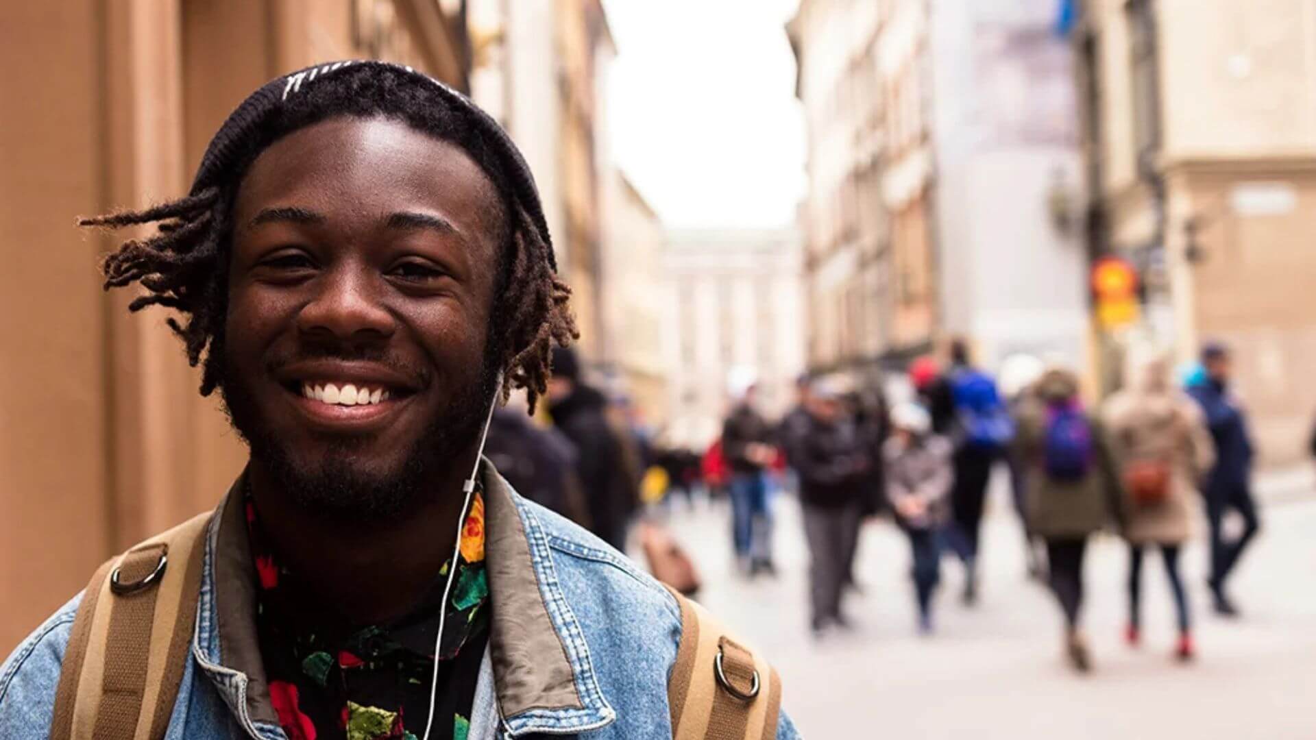 A smiling young man with dreadlocks stands on a bustling city street, surrounded by urban scenery.